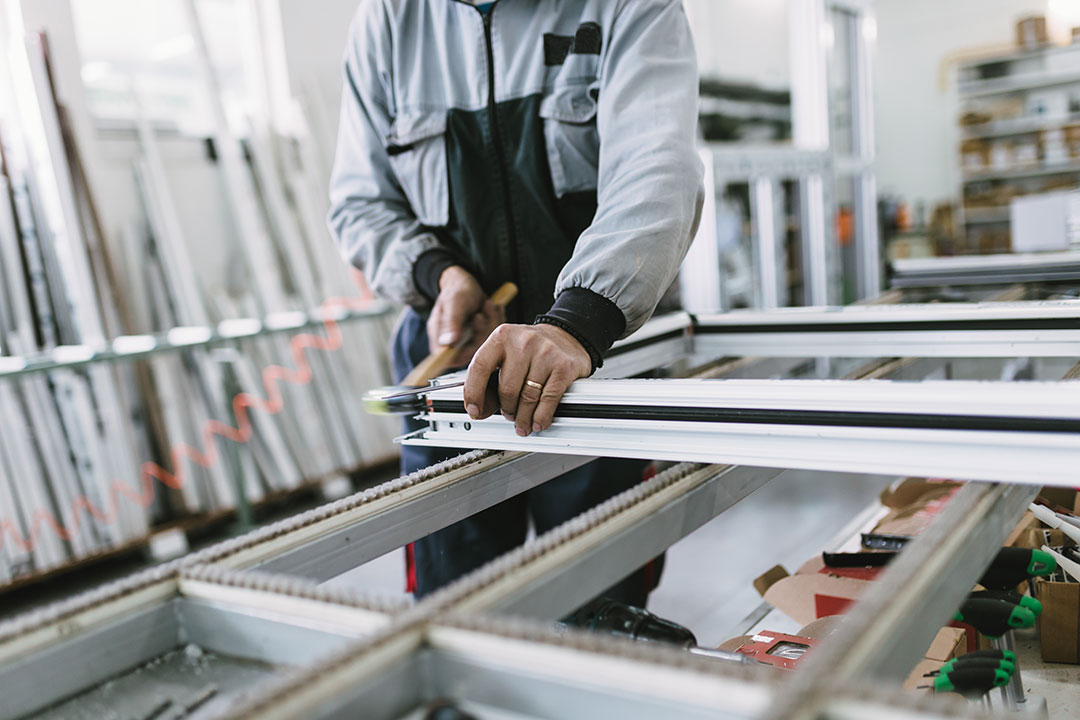 A worker in a light blue uniform assembling a white aluminium window or door frame in a factory setting.